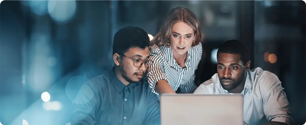 Three colleagues focused on work at a laptop in a dim office.